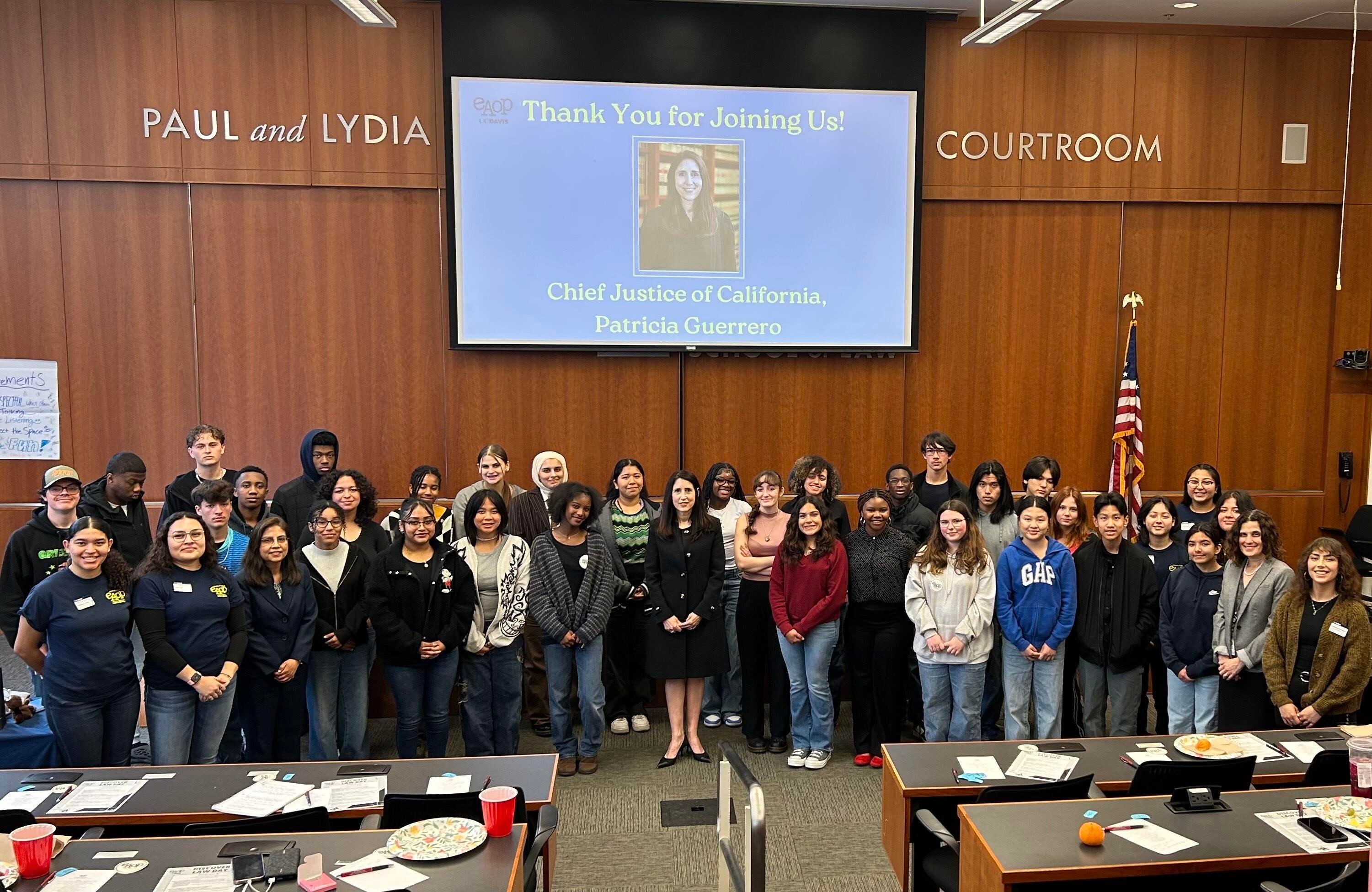 A group of people stand side by side in a courtroom setting beneath a large screen that reads 'Thank You for Joining Us! Chief Justice of California, Patricia Guerrero.'