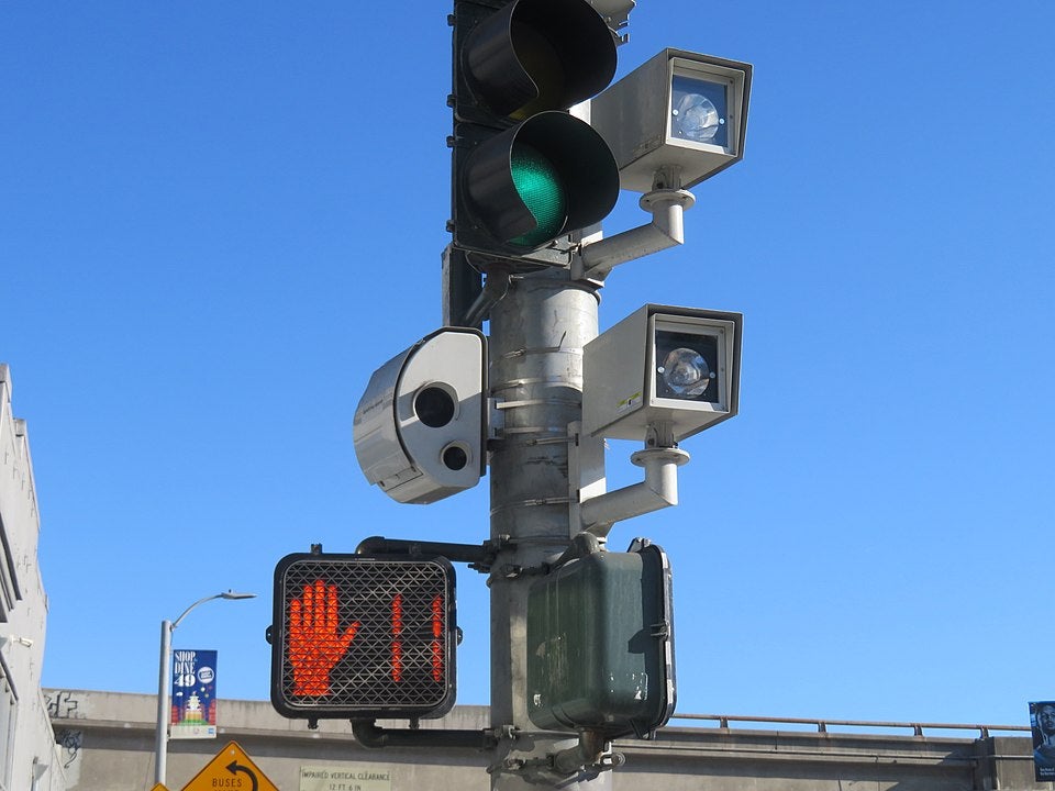 Cameras to record traffic violations are attached to a traffic light pole. A crosswalk sign shows eleven seconds left to cross. 
