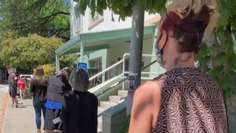 residents stand in line on sidewalk in the sun