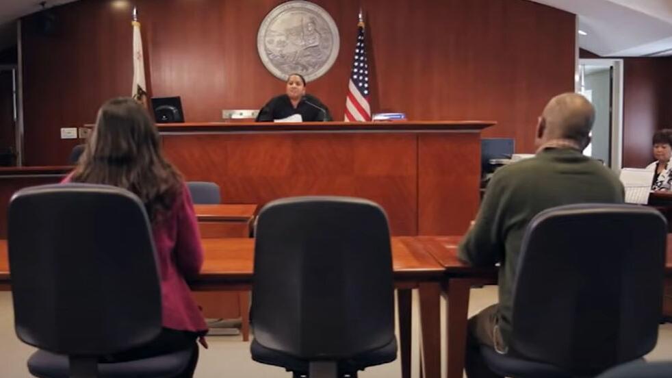 two people sit in a courtroom before a judge on the bench and a court reporter on the side of the room