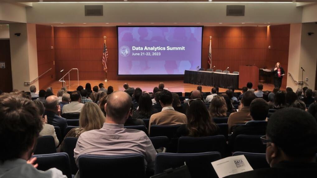 auditorium full of attendees facing stage on screen at front of the room