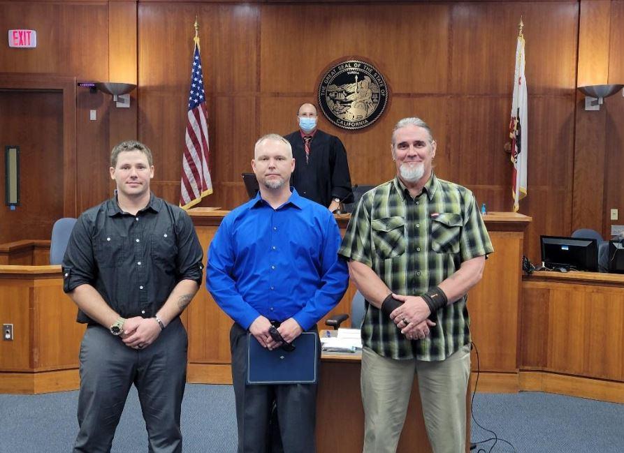 three men stand in front of bench with judge in the background in courtroom