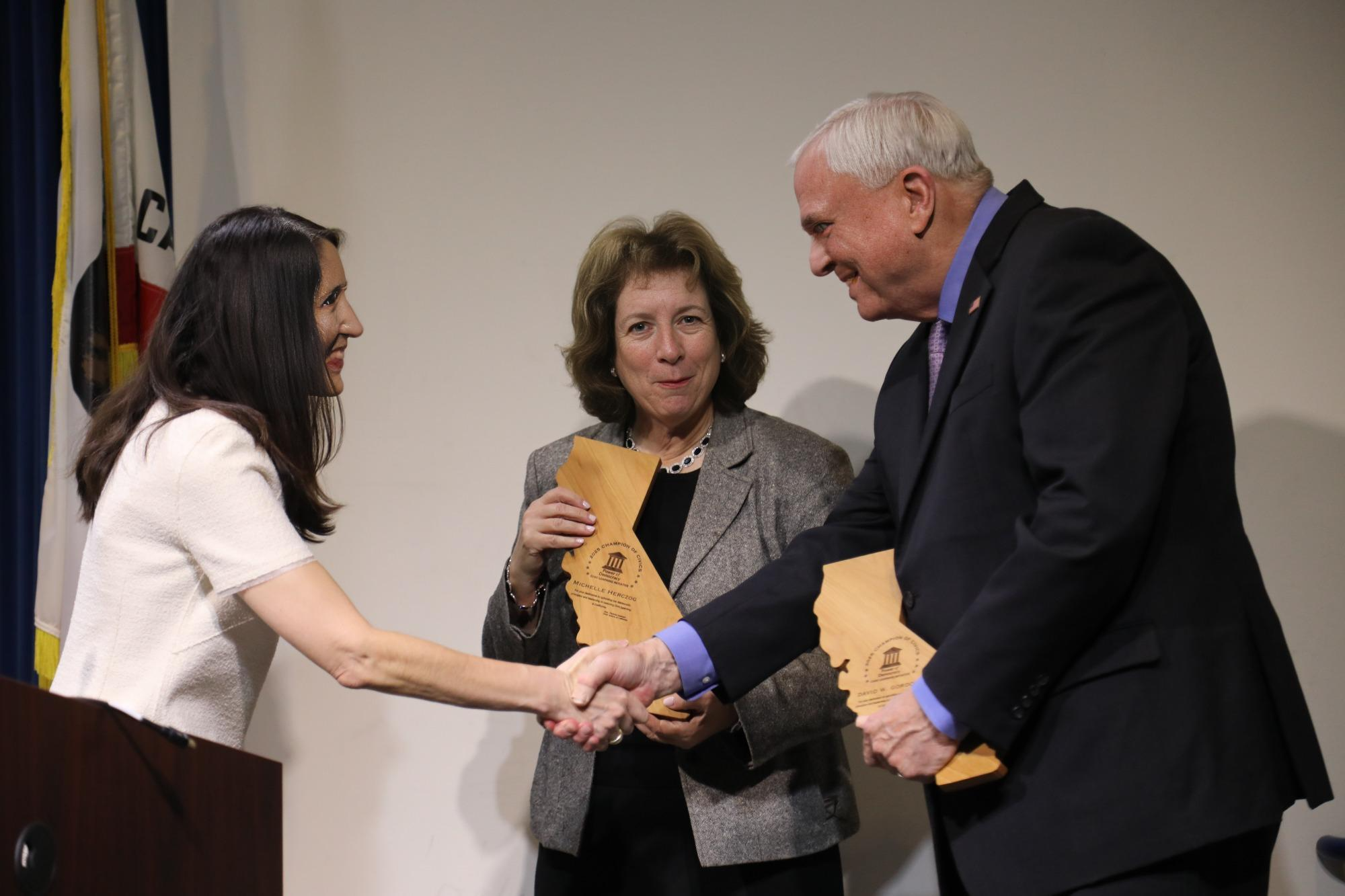 woman shaking hand of man holding wooden award. second woman stands looking at camera holding same award