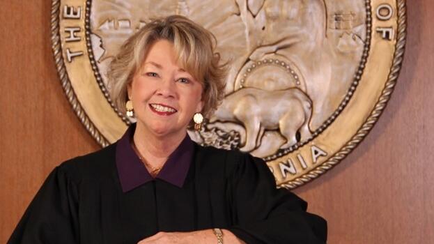 Presiding Justice Kathleen E. O’Leary in judicial attire in front of Seal of the State of California, leaning on brown chair.