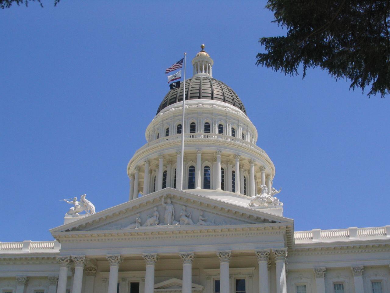 A close up of the California capital building in Sacramento. There are white columns at the front of the building, statues on the roof, and a large dome at the center. 