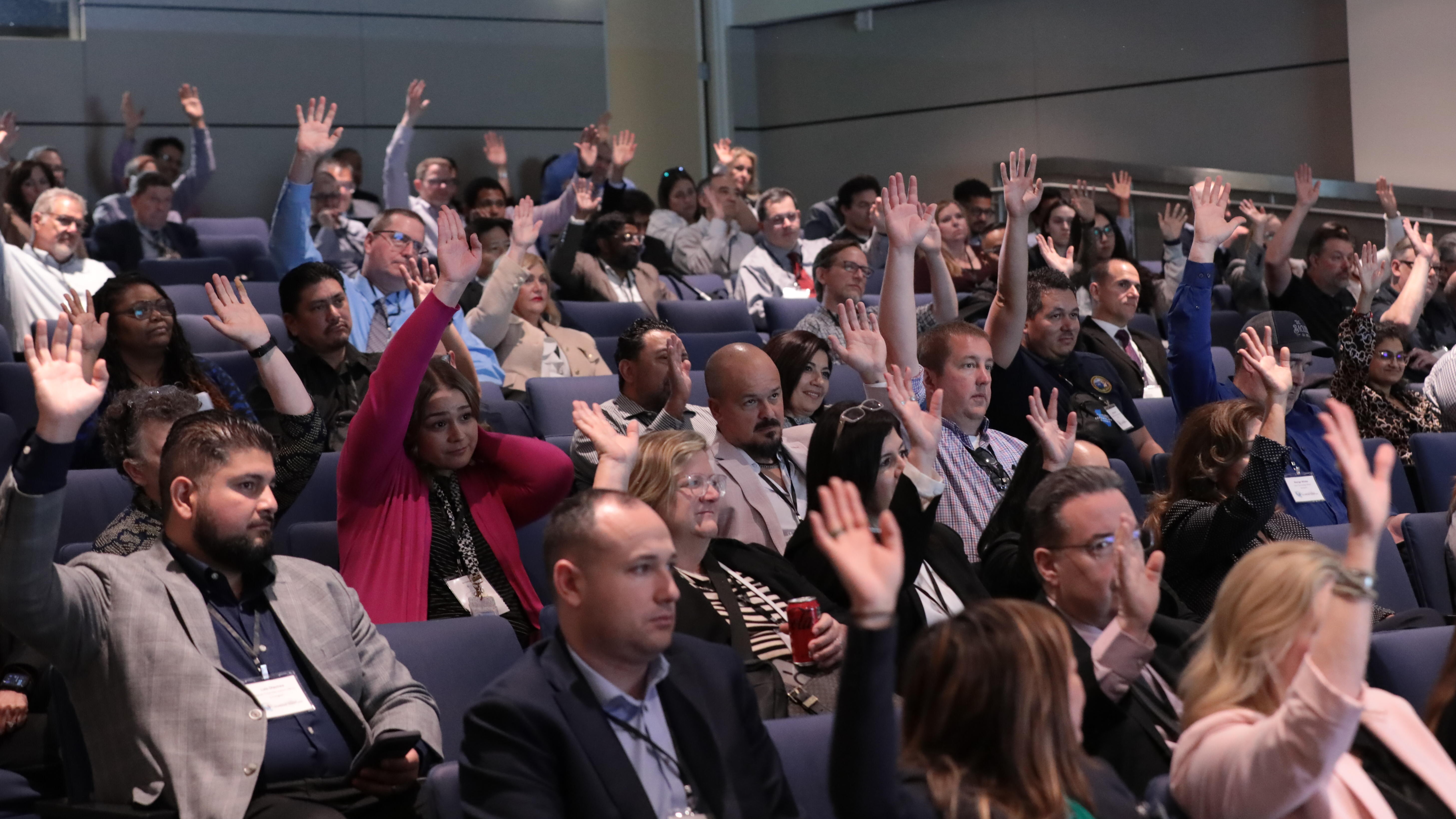 A crowd of people sit in an auditorium raising their hands.