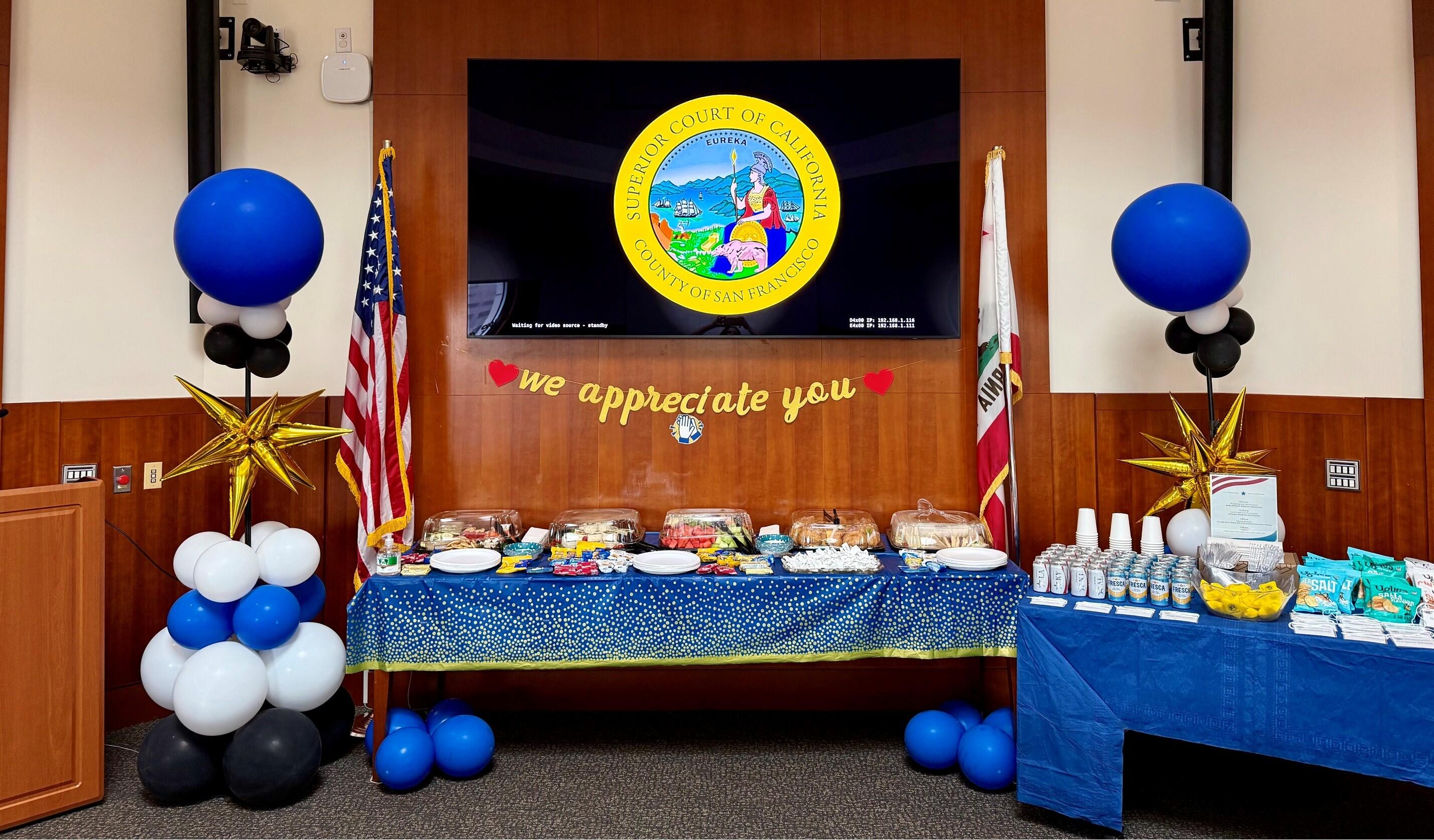 Snacks and drinks sit on two tables with blue tablecloths. Blue and white balloons are next to them. The seal of San Francisco Superior Court is displayed on a monitor. 