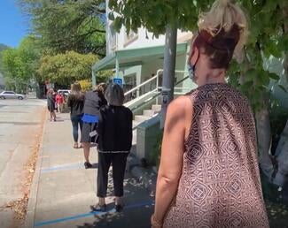 residents stand in line on sidewalk in the sun