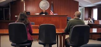 two people sit in a courtroom before a judge on the bench and a court reporter on the side of the room
