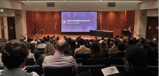 auditorium full of attendees facing stage on screen at front of the room