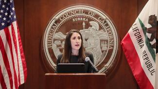 Chief Justice Patricia Guerrero stands at a podium to speak, Judicial Council of California seal behind her, she is flanked by the American flag and the California state flag.