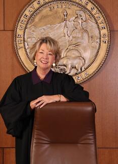 Presiding Justice Kathleen E. O’Leary in judicial attire in front of Seal of the State of California, leaning on brown chair.