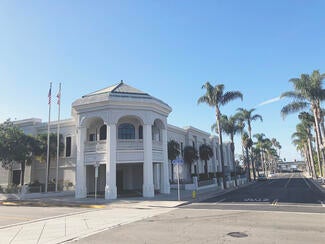 Grey building with columns on a round attached entrance. Palms trees line the road next to it.