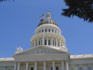 A close up of the California capital building in Sacramento. There are white columns at the front of the building, statues on the roof, and a large dome at the center. 