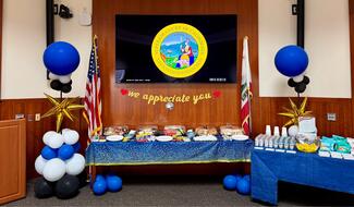 Snacks and drinks sit on two tables with blue tablecloths. Blue and white balloons are next to them. The seal of San Francisco Superior Court is displayed on a monitor. 
