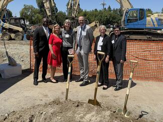 Six people standing in front of a pile of dirt and three shovels at a groundbreaking ceremony
